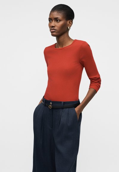 Γυναίκα που φοράει εφαρμοστή πουκαμίσα μακρυμάνικη σε σκουρό πορτοκαλί χρώμα και ψηλόμεσα φαρδιά μπλε παντελόνια με γυναικεία ζώνη μαύρου χρώματος, στέκεται με τα χέρια στις τσέπες.