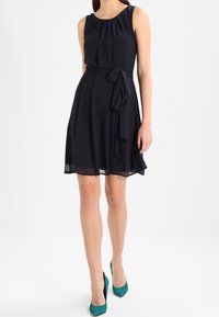 Ärmlös, knälång marinblå klänning i lätt tyg, med en åtsittande midja och knytbälten samt utsvängd kjol. Bärs med turkosa klackar.