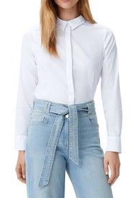 Camicia bianca a maniche lunghe con colletto puntato. Abbinata a jeans a vita alta di colore azzurro chiaro con dettaglio di cintura annodata.