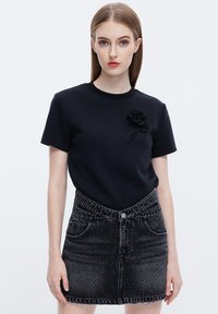 Camiseta de algodón negra con cuello redondo y mangas cortas, que presenta un aplique floral negro texturizado. Combinada con una falda de mezclilla negra desgastada.