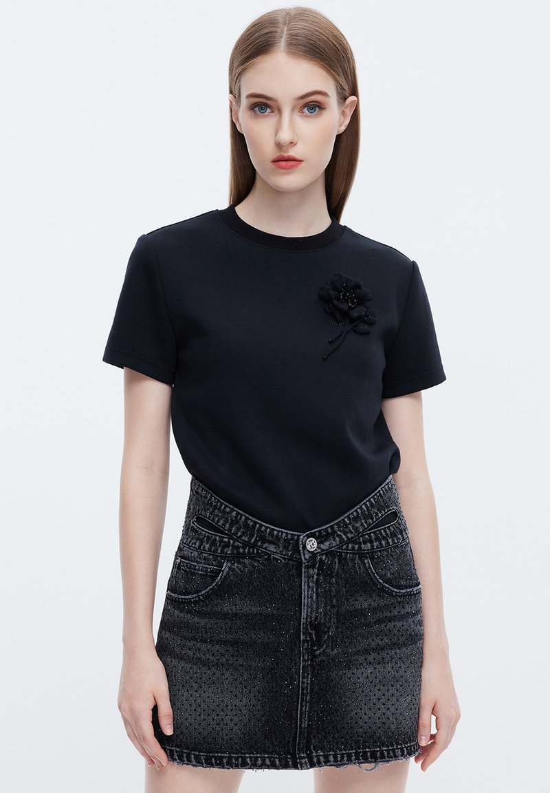 Camiseta de algodón negra con cuello redondo y mangas cortas, que presenta un aplique floral negro texturizado. Combinada con una falda de mezclilla negra desgastada.