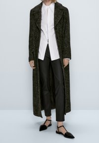 Lång, mörkgrön texturerad kappa med en bred krage, vit skjorta under, svarta åtsittande byxor och svarta spetsiga ballerinas. Ren och skräddarsydd look.