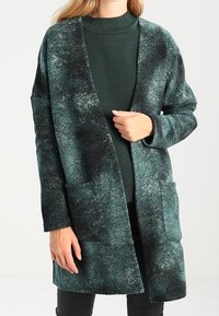 Cardigan lungo verde texturizzato con tasche, caratterizzato da un motivo marmorizzato. Il design aperto aumenta il comfort e la versatilità.