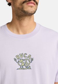 Camiseta de algodón de color púrpura claro con un diseño gráfico que presenta un signo de paz y un globo, acentuado en amarillo y negro, de cuello redondo.