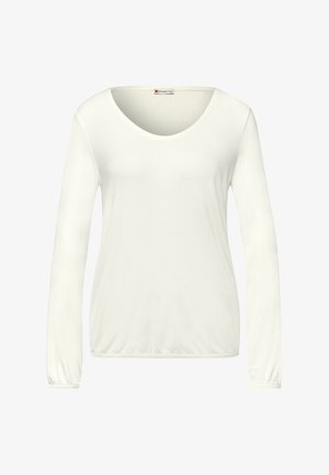 Mujer de cabello largo y oscuro que lleva una camisa de manga larga color crema, pantalones grises con cinturón negro, sosteniendo una chaqueta negra doblada en una mano.