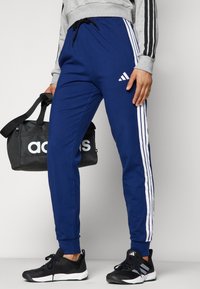 Blå sweatpants i bomullsblandning med vita ränder längs sidorna och en justerbar midja. Svarta sportskor och en svart väska är också synliga.