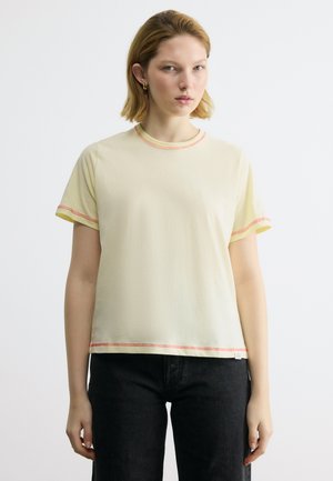 Νεαρή γυναίκα με κοντά ξανθά μαλλιά, που φοράει ένα ανοιχτό κίτρινο κοντομάνικο πουκάμισο με κόκκινες ραφές και μαύρο παντελόνι, στέκεται μπροστά σε λευκό φόντο.