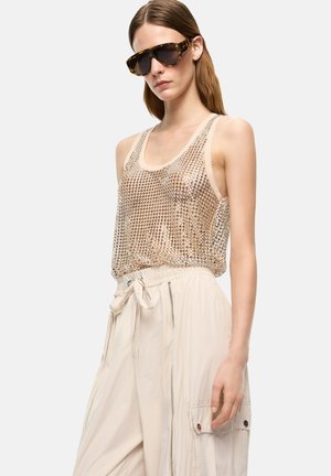 Joven mujer que lleva grandes gafas de sol con montura carey, una camiseta sin mangas de malla metálica y pantalones de cintura alta con cordón en beige claro.