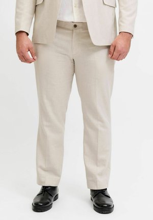 Muž v béžových oblekových nohaviciach, zodpovedajúcom saku, bielej košeli a čiernych leštených spoločenských topánkach stojaci pred jednofarebným pozadím.