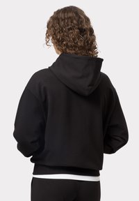 Černá mikina s kapucí, vyrobená z měkké tkaniny, s uvolněným střihem, žebrovanými manžetami a spodním lemem. Hladký povrch bez viditelných vzorů.