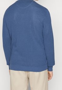 Homme portant un pull à manches longues bleu texturé et un pantalon beige, vu de dos sur un fond uni.