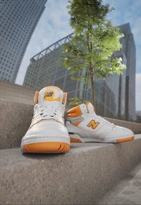 Białe i pomarańczowe sneakersy New Balance o wysokiej cholewce, wykonane z materiałów skórzanych i siatkowych, z widocznym logo oraz pomarańczowymi akcentami na pięcie i podeszwie.