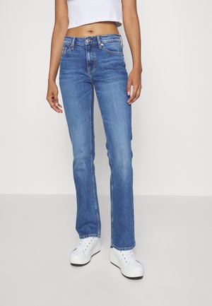 Jeans acampanados de mezclilla azul con un lavado medio, diseño clásico de cinco bolsillos y cierre de botón metálico. Llevados con zapatillas de plataforma blancas.