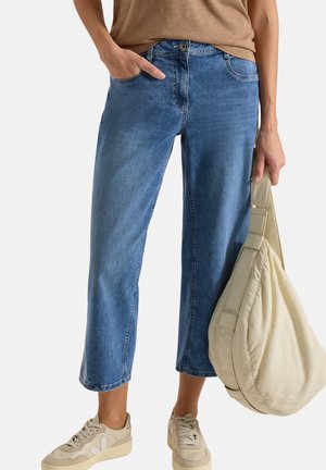 Donna con jeans corti blu, sneakers beige e top marrone, che tiene una grande borsa beige a tracolla nella mano destra.