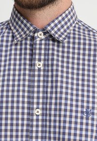 Camicia a quadri blu e grigia con colletto classico, dotata di bottoni bianchi e un logo ricamato sul petto.