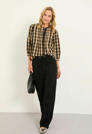 Woman with blonde hair wearing a beige and black checkered blouse, black drawstring pants, silver flats, and holding a black handbag.