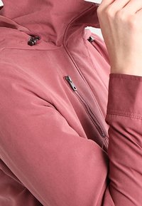 Persona che indossa una giacca color rosa polveroso con una tasca sul petto con cerniera e cappuccio con coulisse, mentre aggiusta il colletto con la mano.
