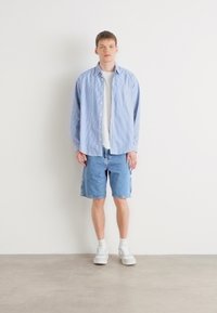 Camisa de botones a rayas azul sobre una camiseta blanca, combinada con pantalones cortos de mezclilla y zapatillas blancas. Fondo neutro, sin patrones ni acentos.