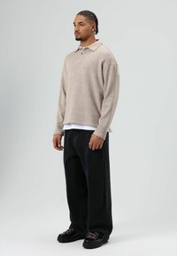 Polo beige à manches longues avec col et un bouton, superposé à une chemise blanche. Associé à un jean noir ample et des chaussures noires.