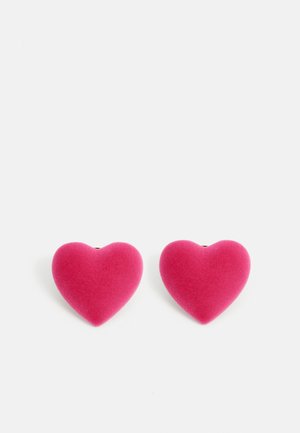 Deux boucles d'oreilles en velours rose en forme de cœur sur un fond blanc, positionnées côte à côte.