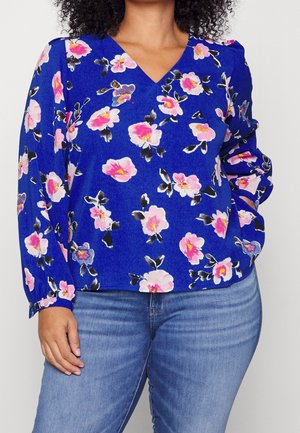 Frau trägt eine blaue Langarmbluse mit pinkem Blumenmuster und blaue Jeans, Hand auf der Hüfte ruhend vor neutralem Hintergrund.