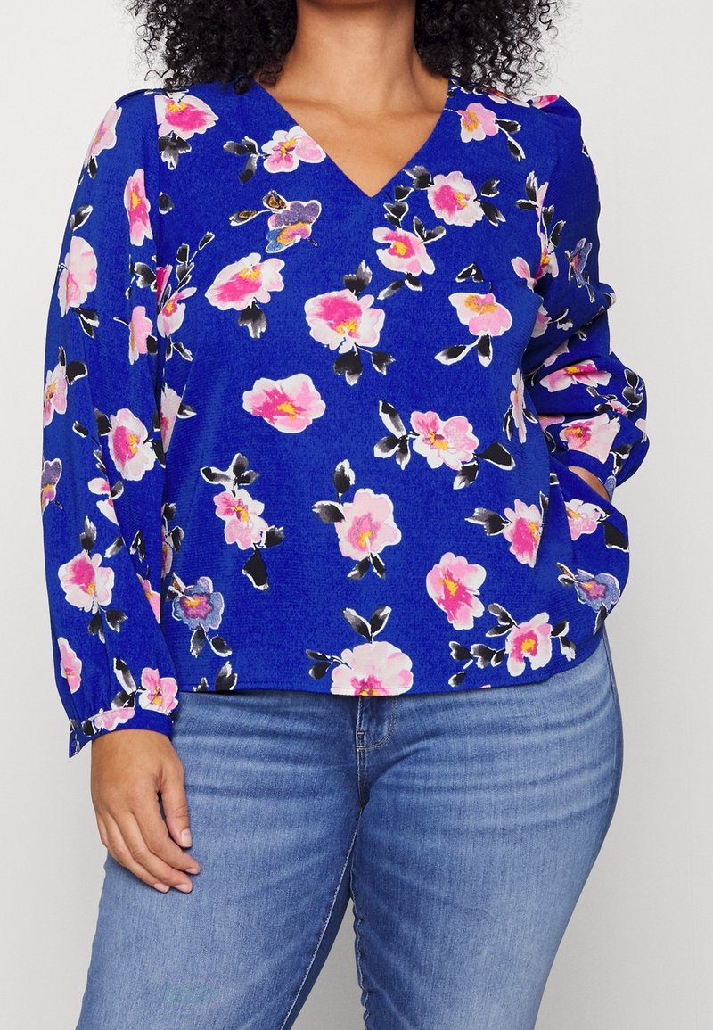 Femme portant un chemisier bleu à manches longues avec imprimé floral rose et un jean bleu, main posée sur la hanche devant un fond uni.