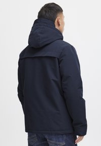Jachetă cu glugă de culoare bleumarin, cu o textură netedă, având un design cu yoke posterior și o croială lejeră, asortată cu blugi din denim închisa.