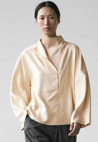 Vrouw die een losse, crèmekleurige blouse draagt met wijde mouwen en twee knopen aan de voorkant, gecombineerd met donkergrijze broeken tegen een neutrale achtergrond.