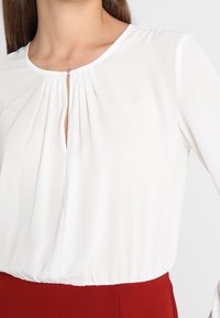 Witte blouse gemaakt van een lichtgewicht stof, met een keyhole halslijn, plooien en een elastische zoom. Gepaard met een rood kledingstuk.