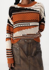 Oranssi, kerma ja musta raidallinen neulepaita, jossa on löysä istuvuus, pyöreä kaula-aukko ja monimutkaiset rei'ityskuvioinnit. Yhdistetty ruskeaan nahkahameeseen.