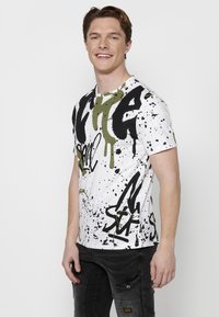 Camiseta blanca de manga corta con diseños de estilo graffiti en negro y verde aceituna, con acentos de salpicaduras a lo largo y un escote redondo.