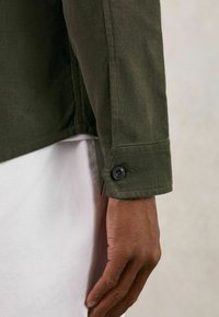 Olivgrüne Cordjacke mit einem geknöpften Bündchen. Der Stoff hat eine gerippte Textur und zeichnet sich durch ein einfaches Design aus. Darunter ist ein weißes Hemd sichtbar.