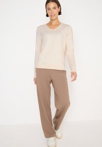 Helle beige Langarmbluse mit V-Ausschnitt, kombiniert mit lockeren braunen Hosen; das Model trägt weiße und braune Sneakers. Einfaches, lässiges Design.