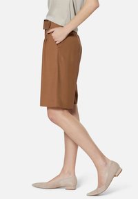 Bruine op maat gemaakte shorts met zijzakken en een stoffen riem, gecombineerd met lichtbeige suède platte schoenen, weergegeven vanuit een zijaanzicht.
