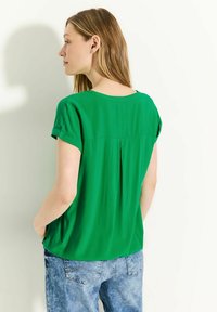 Blusa verde a maniche corte con una vestibilità leggermente ampia, caratterizzata da una piega sul retro. Abbinata a jeans in denim di un leggero blu.