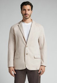 Blazer de lino beige claro con dos botones, solapas de muesca y dos bolsillos frontales, llevado sobre una camiseta blanca de cuello redondo. Textura suave.