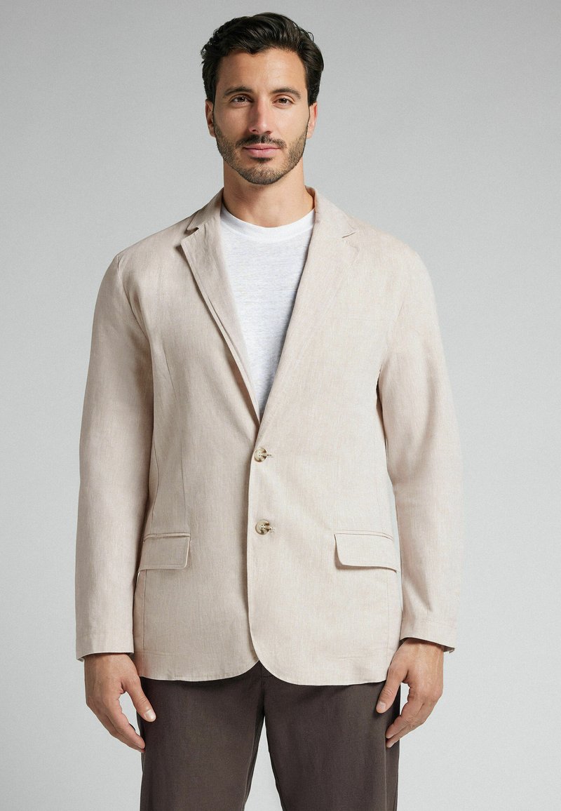Blazer de lino beige claro con dos botones, solapas de muesca y dos bolsillos frontales, llevado sobre una camiseta blanca de cuello redondo. Textura suave.