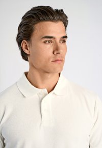 Vit pikétröja i mjukt tyg med krage och tre knappar vid halsen; slät textur och figurnära design.