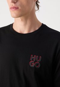 Zwarte katoenen t-shirt met een ronde halslijn en een gestructureerd rood en zwart "HU GO" logo aan de linkerkant van de borst.