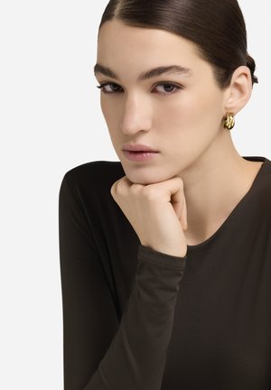 Zlatna naušnica u obliku obruča s isprepletenim dizajnom, nošena od strane osobe u uskoj, dugoj tamnosmeđoj majici. Vidljiva je glatka tekstura kože.