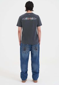 Camiseta gris con gráficos de flores y alas en la parte de atrás, combinada con jeans azul que tienen telarañas bordadas y acentos negros.