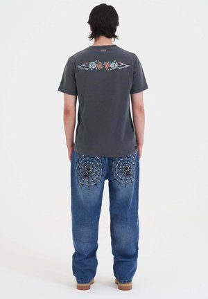 T-shirt grigia con grafiche floreali e ali sul retro, abbinata a jeans blu con ragnatele ricamate e dettagli neri.