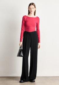 Ženska stoji oblečena v oprijet rdeč dolg rokav top, široke črne hlače, črne čevlje in drži majhno črno torbico.