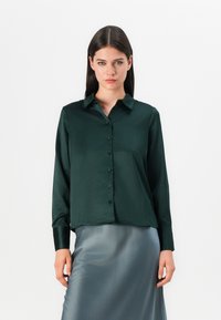 Camicia in satin verde scuro con bottoni, colletto, maniche lunghe e orlo dritto. Indossata con una gonna liscia e lucida grigia.