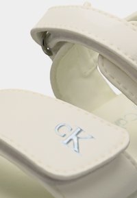 Κοντινή λήψη λουριού σανδαλιού εκρού με μεταλλική αγκράφα και λογότυπο "cK" ανάγλυφο στο μπροστινό λουρί.