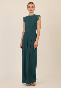 Teal jumpsuit met een kanten lijfje, kapmouwen, hoge hals en wijde broekspijpen. Heeft plooien en zilveren schoenen. Gladde textuur.