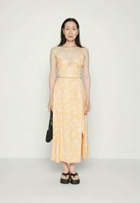 Mulher num vestido midi amarelo com estampado floral e alças finas, sandálias de plataforma pretas, segurando uma bolsa preta contra um fundo plain.