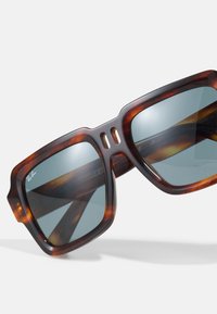 Ochelari de soare tortoiseshell maro cu rame mari, în formă pătrată și lentile gradient. Caracteristicile includ detalii tăiate și un finisaj lucios.
