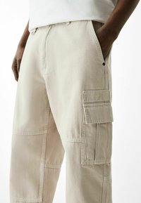 Beiga cargobyxor gjorda av slitstarkt tyg, med stora sidofickor, bälteslinor och en avslappnad passform. Synlig sömnadsdetalj överallt.