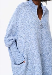 Person som bär en mjuk, ljusblå stickad zip-up tröja med ringdragkedja och en hand i fickan, visad från halsen till midjan.